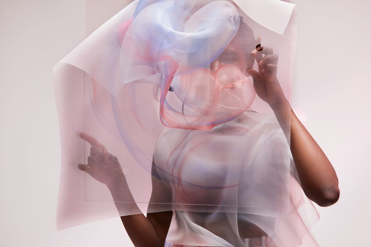 A person stands behind or partially wrapped in layers of translucent fabric in soft pink, blue, and red tones. The material flows gently, creating a dreamy, abstract effect. The person's silhouette is visible through the fabric, with one hand raised and touching it. The interplay of light, color, and texture gives the scene a surreal, artistic atmosphere.