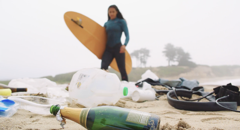 Image of trash on a beach in focus and a surfer in the background